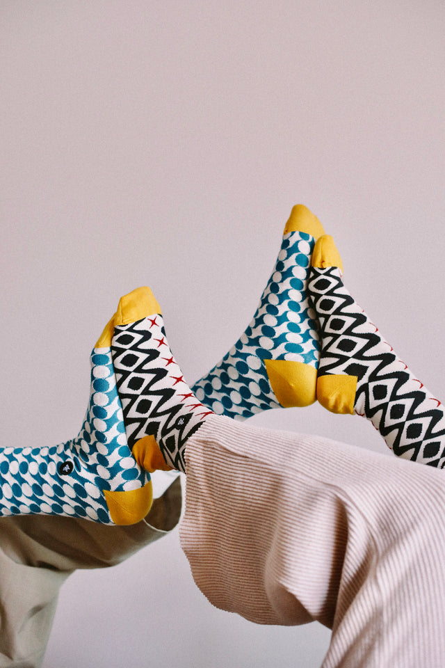 Two persons' legs, up in the air with their feet pressed together, one wearing black geometric patterned socks and the other wearing petrol geometric patterned socks, against a light grey background.