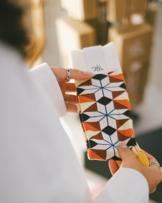 A pair of colourful geometric socks is contemplated in the hands of a woman wearing a white shirt.