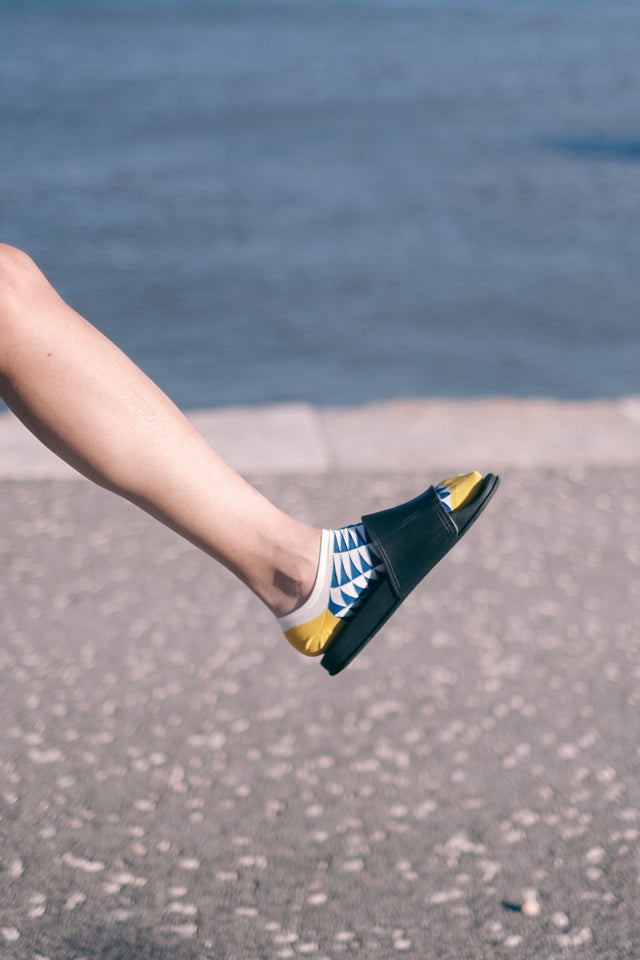 A woman's leg wearing black sandals and blue geometric ankle socks with yellow toe and heels, with a riverside background.