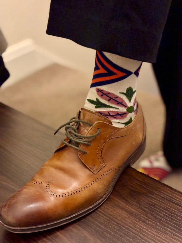 A man’s foot over a bench wearing brown Oxford shoes and patterned socks featuring purple and orange diamond shapes and pink flowers with green leaves, standing on a beige floor.