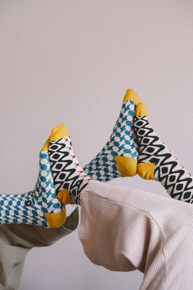 Two persons' legs, up in the air with their feet pressed together, one wearing black geometric patterned socks and the other wearing petrol geometric patterned socks, against a light grey background.