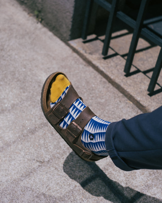 A foot adorned with blue geometric socks featuring a yellow toe cap peeks out from sandals, complemented by blue trousers, as it strolls along a sunlit street.
