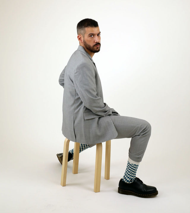 A man sitting on a wooden bench, wearing a light grey suit and black shoes, looks at the camera over his right shoulder, while letting the geometric blue pattern of his socks show.