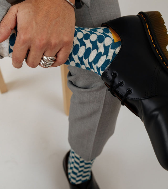 On a wooden bench, a man's legs are visible, dressed in a light grey suit. His left hand crosses over his right leg, revealing the blue geometric pattern of his socks.