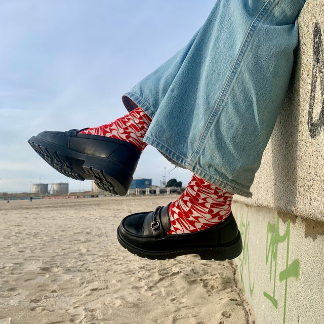 Legs of a person sitting on a stone wall, showing a pair of tiled red socks and black shoes, while contemplating the sunset by the sea.