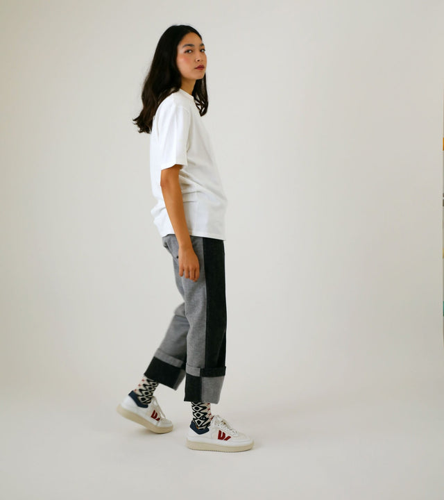 A standing woman wears a white T-Shirt paired with rolled-up bicolour jeans, showcasing her black geometric patterned socks. White trainers adorn her feet as her arms rest along her body.