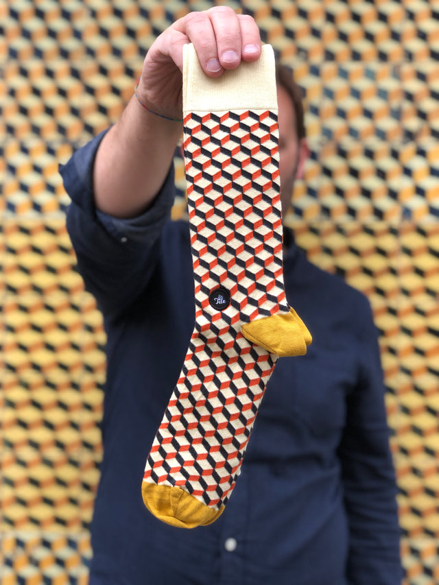 A man showcasing a pair of geometric patterned socks, holding them up in front of a wall with a matching design.