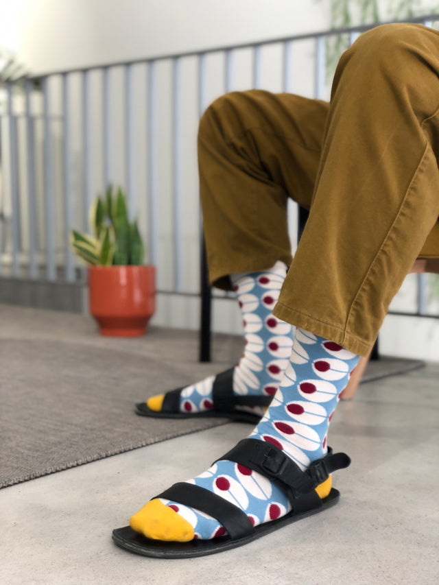 Someone wearing mustard-colored pants and Olhão socks with blue dots and red circles, paired with black sandals. They are sitting indoors near a plant in an orange pot.