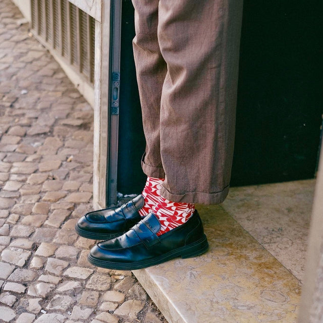 A person standing at the entrance of a building, wearing brown trousers and black penny loafers, revealing geometric red patterned socks.