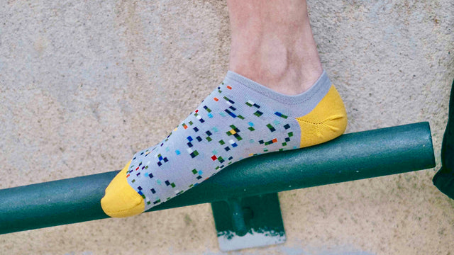 A person's foot, wearing polychromatic ankle socks with a grey base, yellow toe caps, and heels, rests on a metal handrail fixed to an outdoor wall.