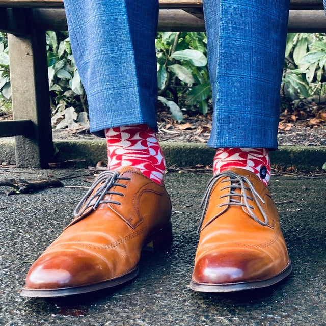 The legs of a man are visible as he sits on a park bench, dressed in a blue suit and brown Oxford shoes. He allows the geometric red pattern of his socks to peek out.