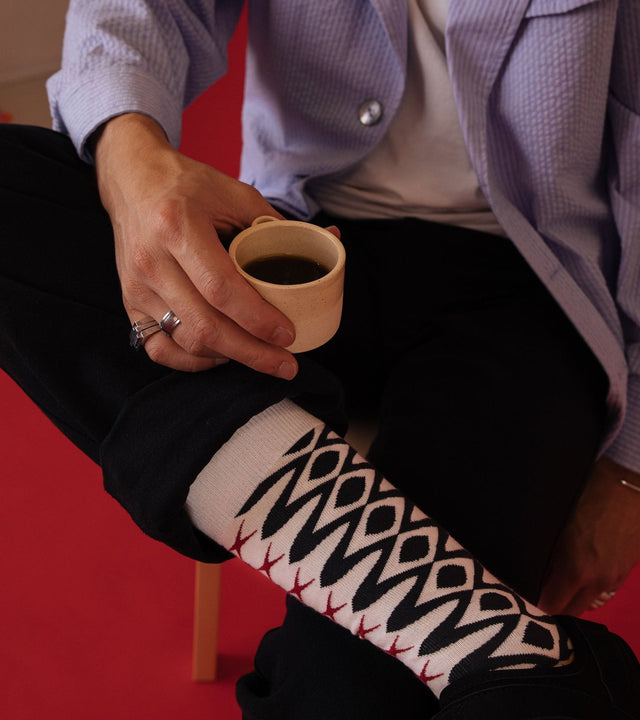 A man sits with one leg crossed over the other, dressed in a light blue jacket and black trousers. He holds a cup of coffee, showcasing his bold black patterned socks.