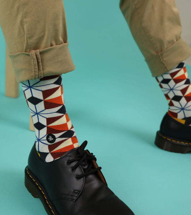 A close-up of a man's legs as he sits on a wooden bench, wearing rolled-up khaki trousers. He proudly displays his geometric patterned socks, paired with black shoes.